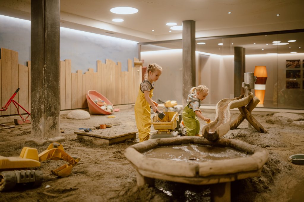 Zwei Kinder spielen in einem Sandbereich mit Wasser und Spielzeug in einem Innenraum mit Säulen und gedämpfter Beleuchtung