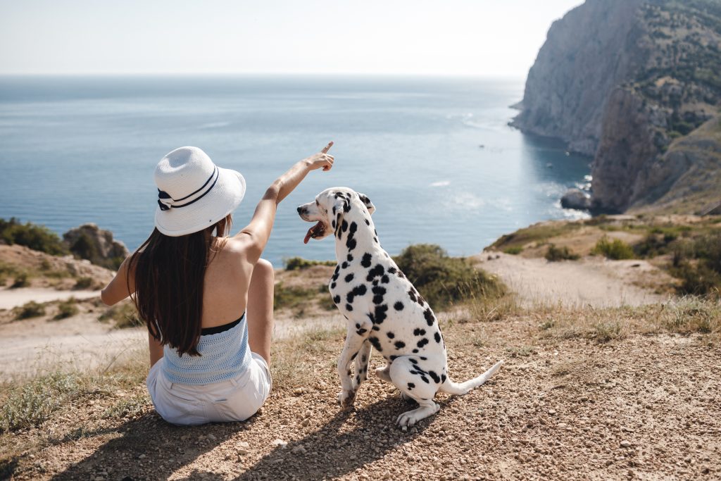 Frau mit Sonnenhut sitzt neben Dalmatiner auf einem Felsen und zeigt aufs Meer mit Klippen im Hintergrund