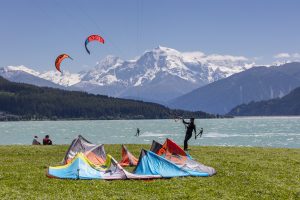 Kitesurfer am See mit mehreren auf dem Gras liegenden bunten Kites und schneebedeckten Bergen im Hintergrund