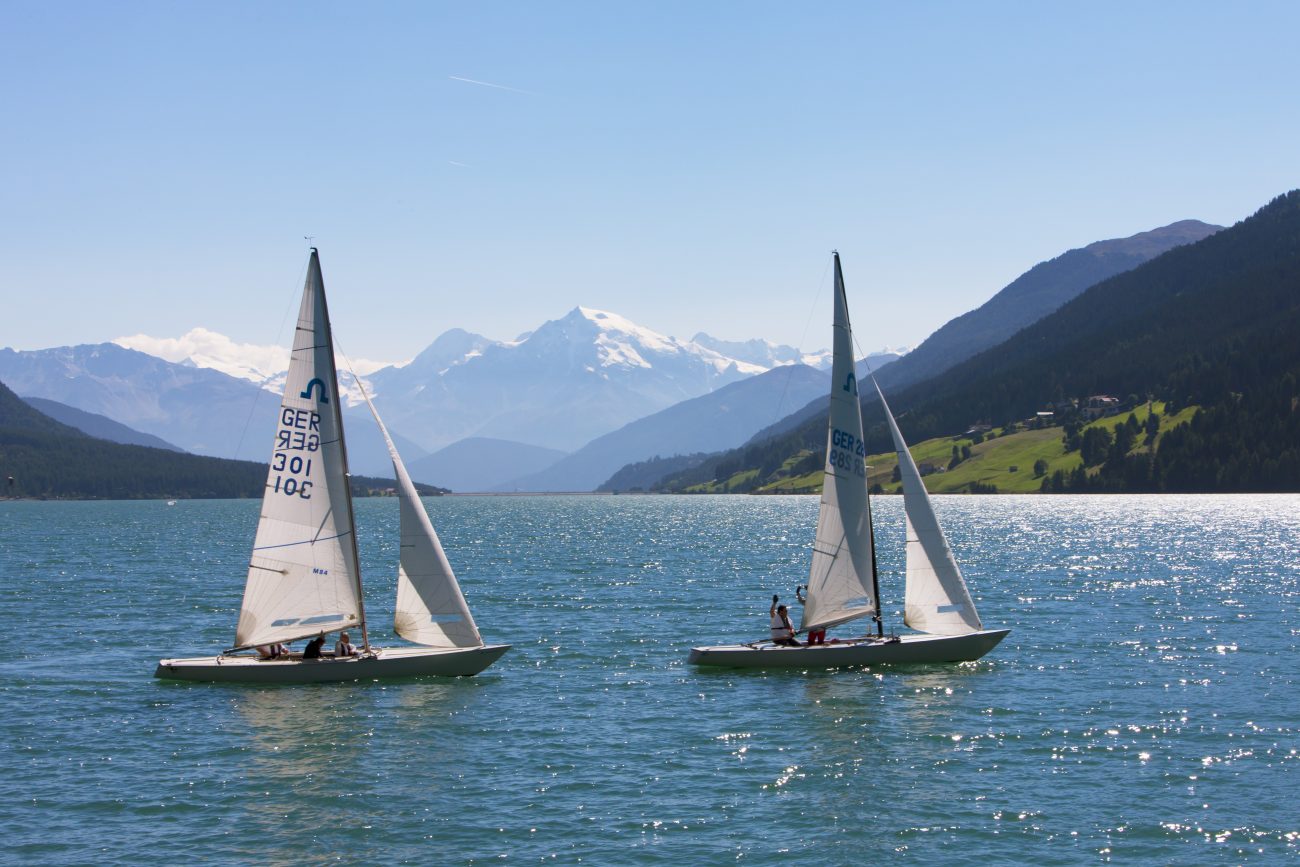 Zwei Segelboote mit weißen Segeln auf einem See vor Bergkulisse bei klarem Himmel