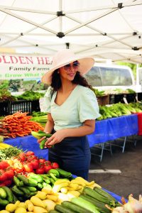 Nisha Vora mit Sonnenhut und Sonnenbrille steht an einem Marktstand mit verschiedenen Gemüsesorten wie Tomaten, Gurken und Zucchini