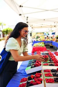 Nisha Vora mit lockigem Haar und Sonnenbrille greift an einem Marktstand mit verschiedenen Beeren in kleinen Schalen