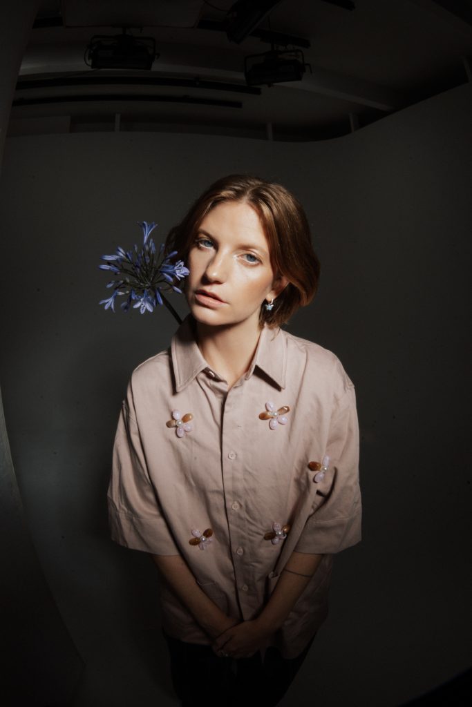 Wilhelmine mit kurzem braunem Haar trägt helles Hemd mit Blütenapplikationen und hält blaue Blume hinter dem Kopf