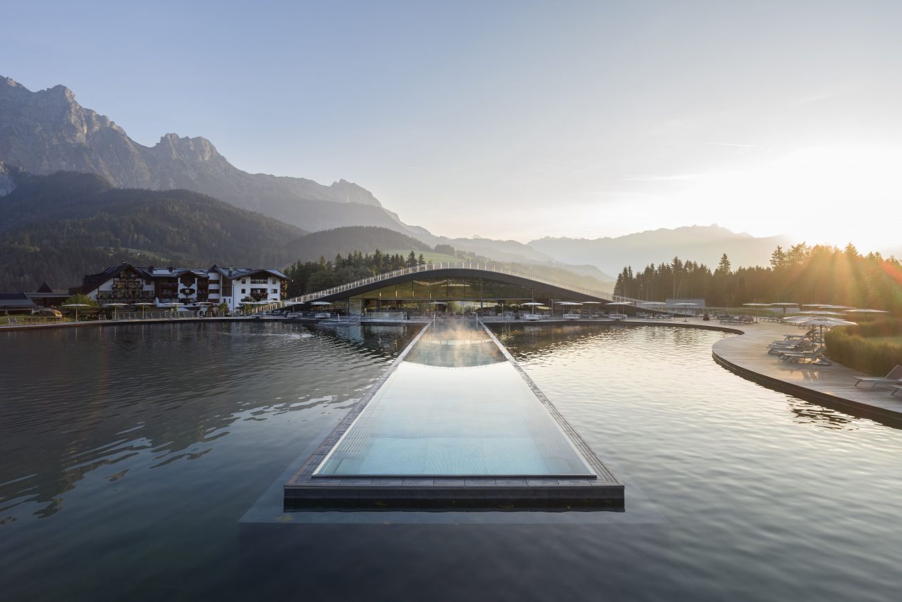 Außenansicht eines Hotels mit Brücke über einen See und Bergkulisse im Sonnenaufgang