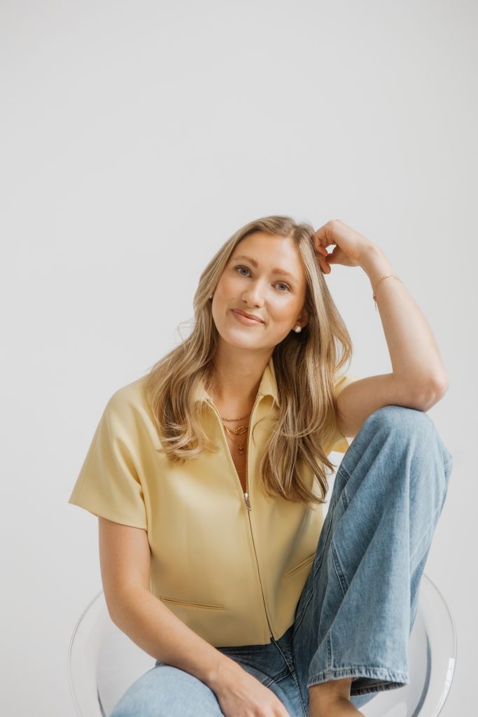 Luisa Werner mit langen blonden Haaren sitzt lächelnd mit einem angewinkelten Bein auf einem transparenten Stuhl und trägt ein hellgelbes Hemd und blaue Jeans