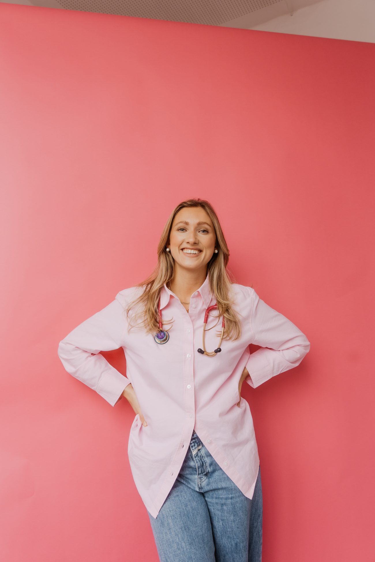 Luisa Werner mit langem Haar, weißem Kittel und Stethoskop vor rosa Hintergrund, die lächelnd die Hände in die Hüften stemmt