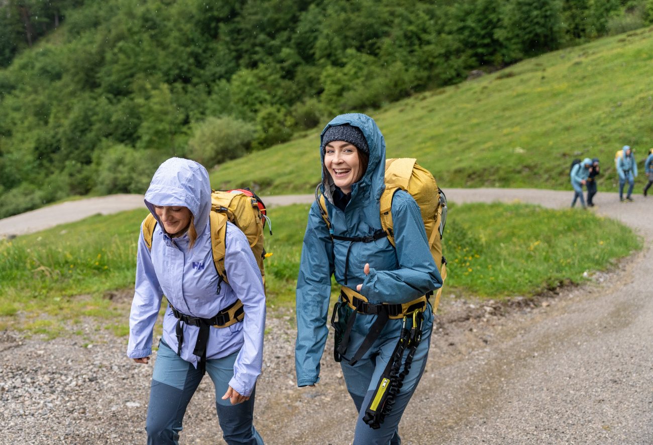 Zwei Wanderinnen mit Rucksäcken und Regenjacken gehen einen Schotterweg bergauf, im Hintergrund weitere Wanderer auf einem grünen Hang