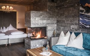 Wohnzimmer mit blauem Sofa, Kamin aus grauem Stein, Holzbalkendecke und Doppelbett im Hintergrund