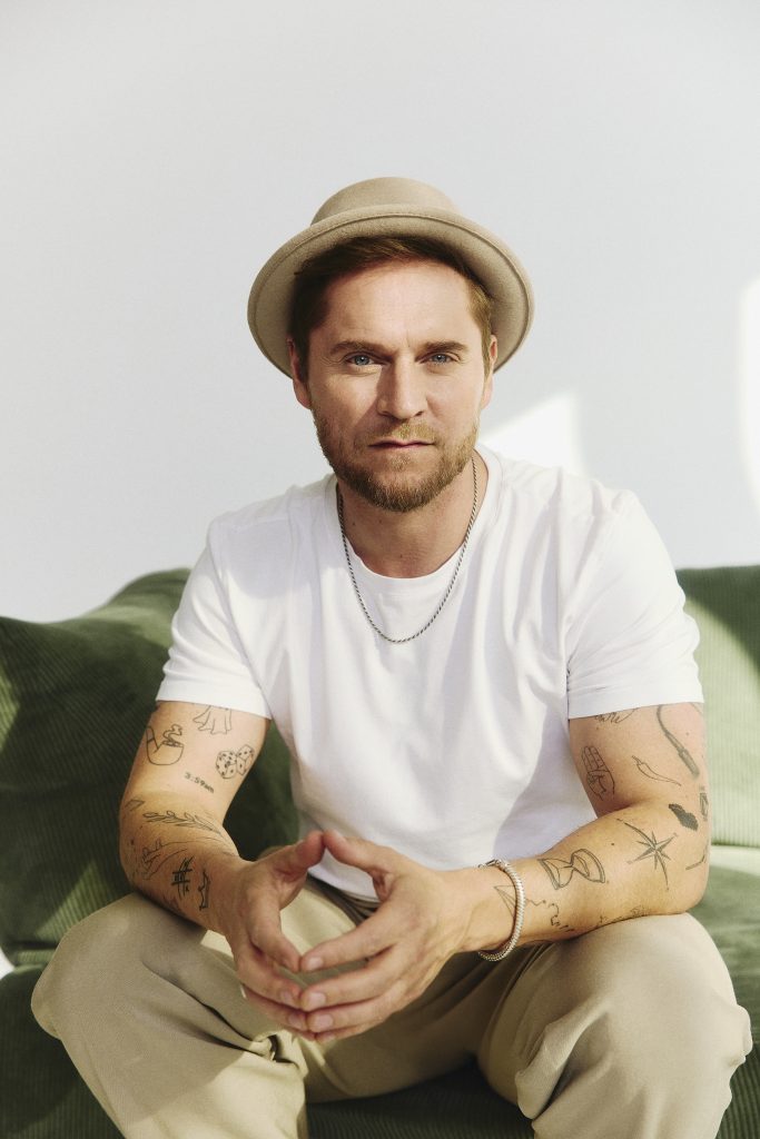 Johannes Oerding mit Hut, weißem T-Shirt und Tattoos an den Armen sitzt auf grünem Sofa und schaut frontal in die Kamera