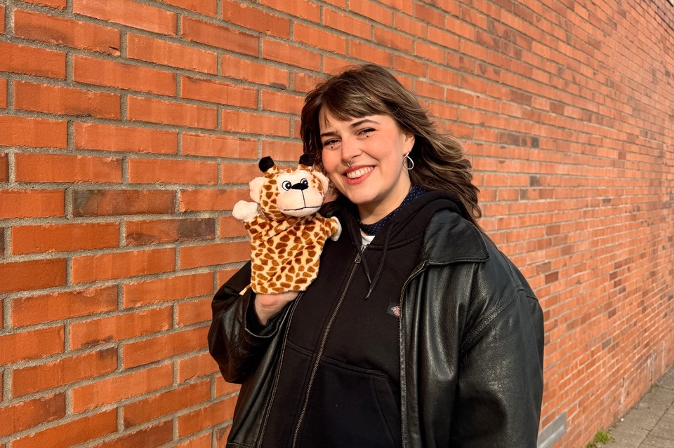 SOFFIE mit schwarzer Lederjacke und blauer Jeans hält ein Giraffen-Handpuppe vor einer roten Backsteinmauer und lächelt