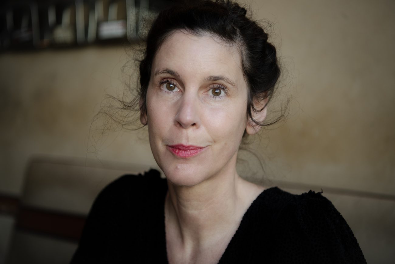 Daniela Dröscher mit zusammengebundenem dunklem Haar, schwarzem Oberteil und neutralem Gesichtsausdruck vor unscharfem Hintergrund