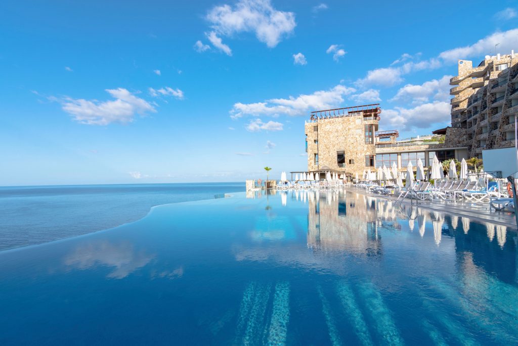 Infinity-Pool mit Blick auf das Meer und angrenzendem Hotelgebäude unter blauem Himmel mit Wolken