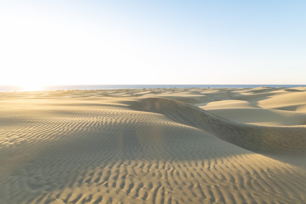 Weite Sanddünenlandschaft unter hellem Himmel mit Sonnenlicht am Horizont