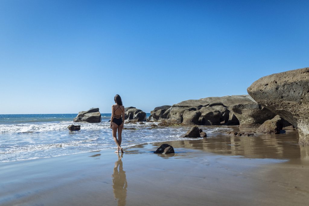 Frau in Badebekleidung geht am Strand mit Felsen im Hintergrund entlang
