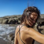 Frau mit offenem Haar und schwarzem Trägerkleid blickt seitlich am felsigen Strand mit Meer im Hintergrund