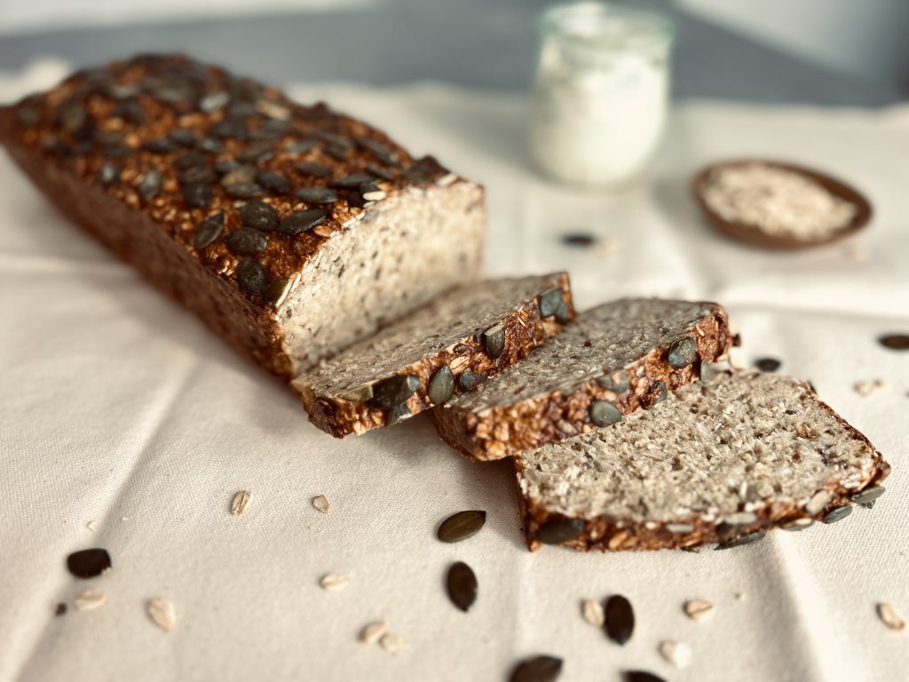 Dunkles Brot mit Haferflocken und Kürbiskernen, drei Scheiben liegen auf einem weißen Tuch, im Hintergrund ein Glas mit Quark und eine Schale mit Haferflocken