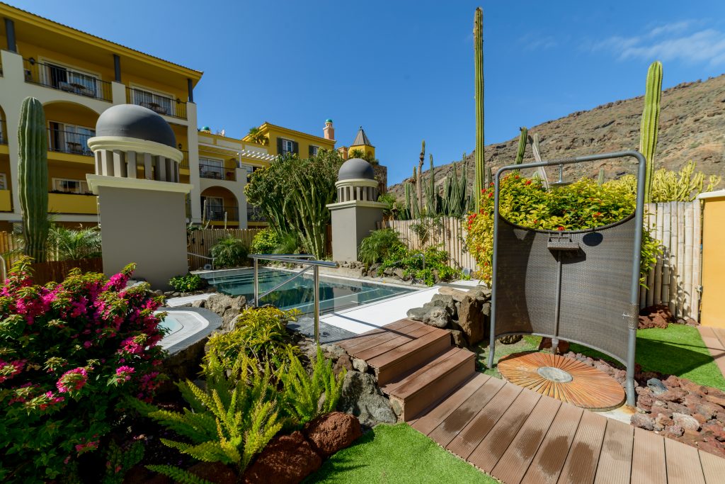 Hotelgarten mit Pool, Holzsteg, exotischen Pflanzen und Kakteen vor einem gelben Gebäude und Bergkulisse