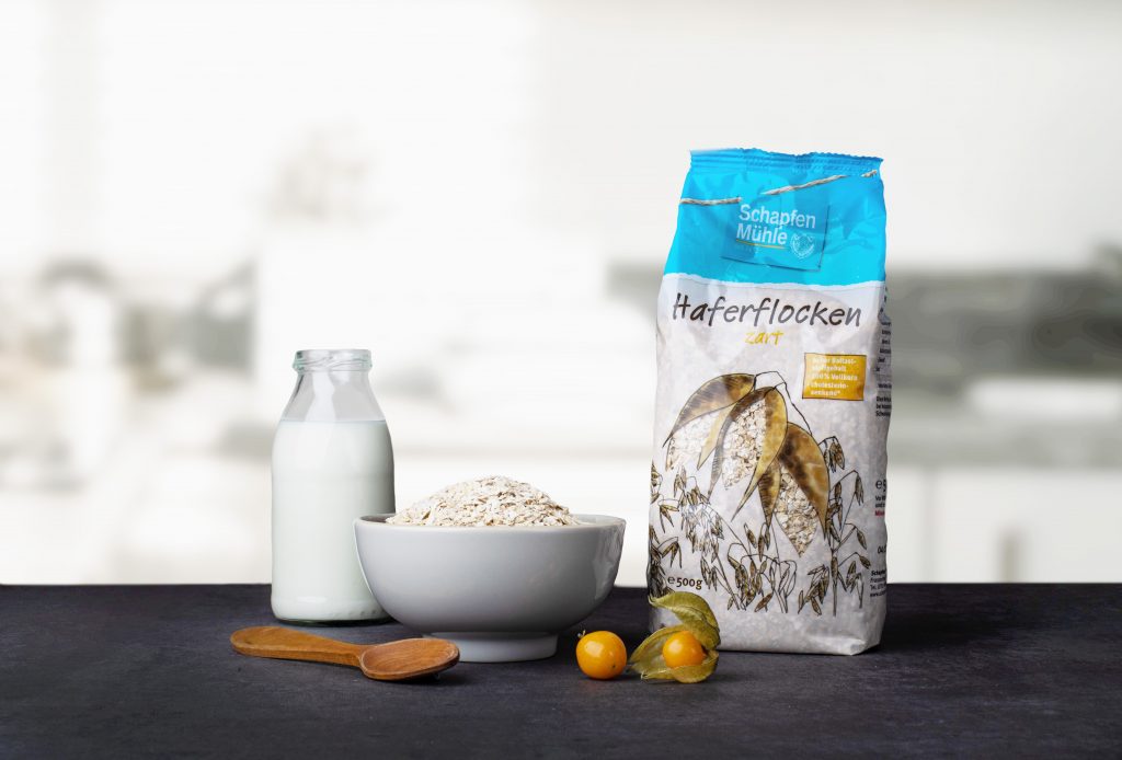 Schale mit Haferflocken, Glasflasche mit Milch, Holzlöffel, zwei Physalis und Packung Haferflocken der Marke Schapfen Mühle auf dunklem Untergrund