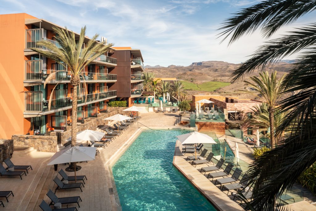 Hotelanlage mit langem Pool, Liegestühlen, Sonnenschirmen und Palmen vor bergiger Landschaft