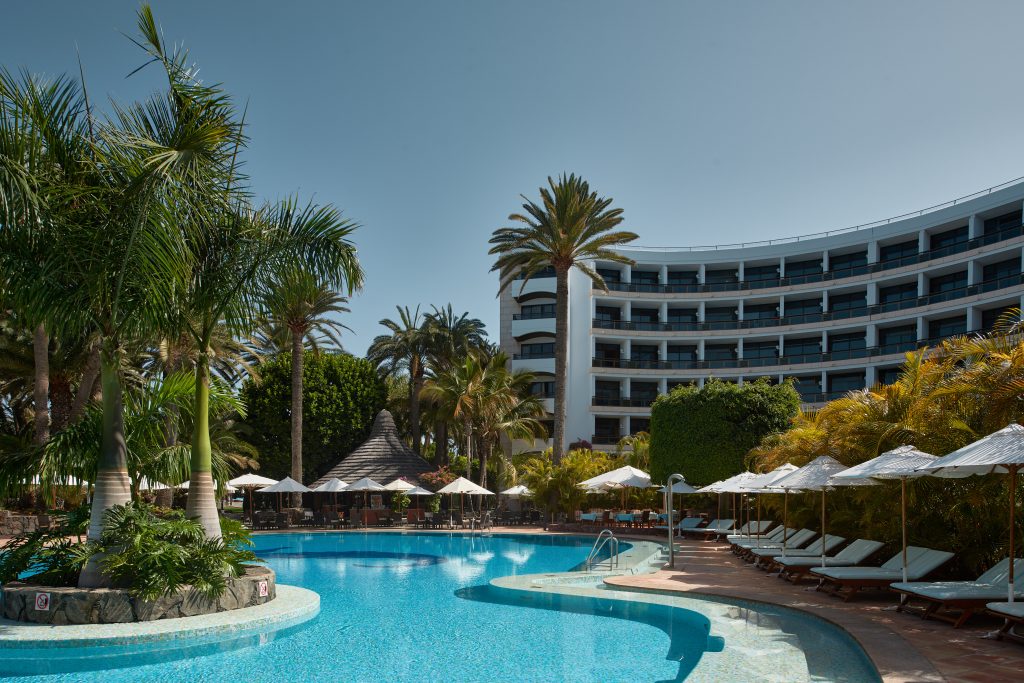 Hotel mit geschwungenem mehrstöckigem Gebäude, umgeben von Palmen und einem Pool mit Liegestühlen und Sonnenschirmen