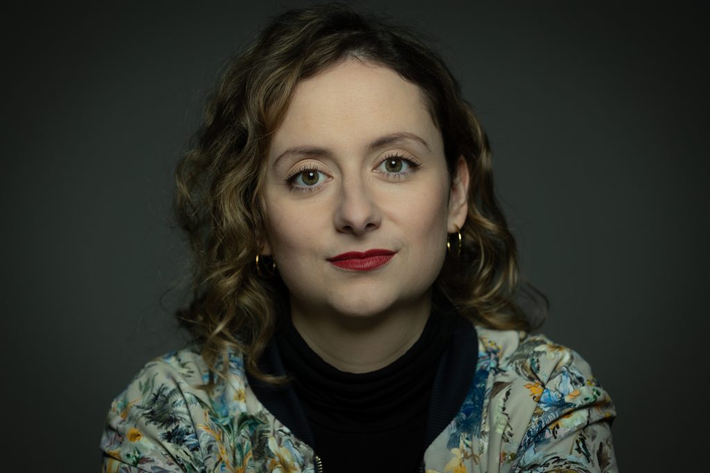 Alexandra Zykunov mit lockigem Haar, rotem Lippenstift und floral gemusterter Jacke vor dunklem Hintergrund