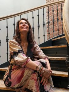 Dr. Petra Algenstaedt sitzt auf einer Treppenstufe, schaut lächelnd nach oben, trägt gemustertes Kleid und mehrere Armbänder