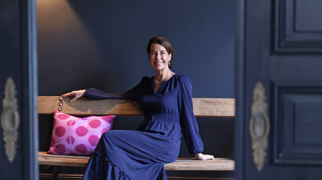 Dr. Petra Algenstaedt in blauem Kleid sitzt lächelnd auf einer Holzbank mit pinkem Kissen vor dunkler Wand