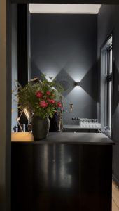 Dunkler Empfangstresen mit großer Vase voller roter Blumen und grüner Zweige vor dunkelgrauer Wand mit indirekter Beleuchtung und Fenster rechts