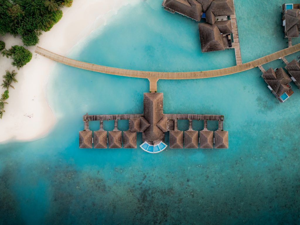 Luftaufnahme eines Stegs mit mehreren über Wasser gebauten Bungalows und einem Pool am Ende des Stegs in türkisfarbenem Wasser neben einem weißen Sandstrand