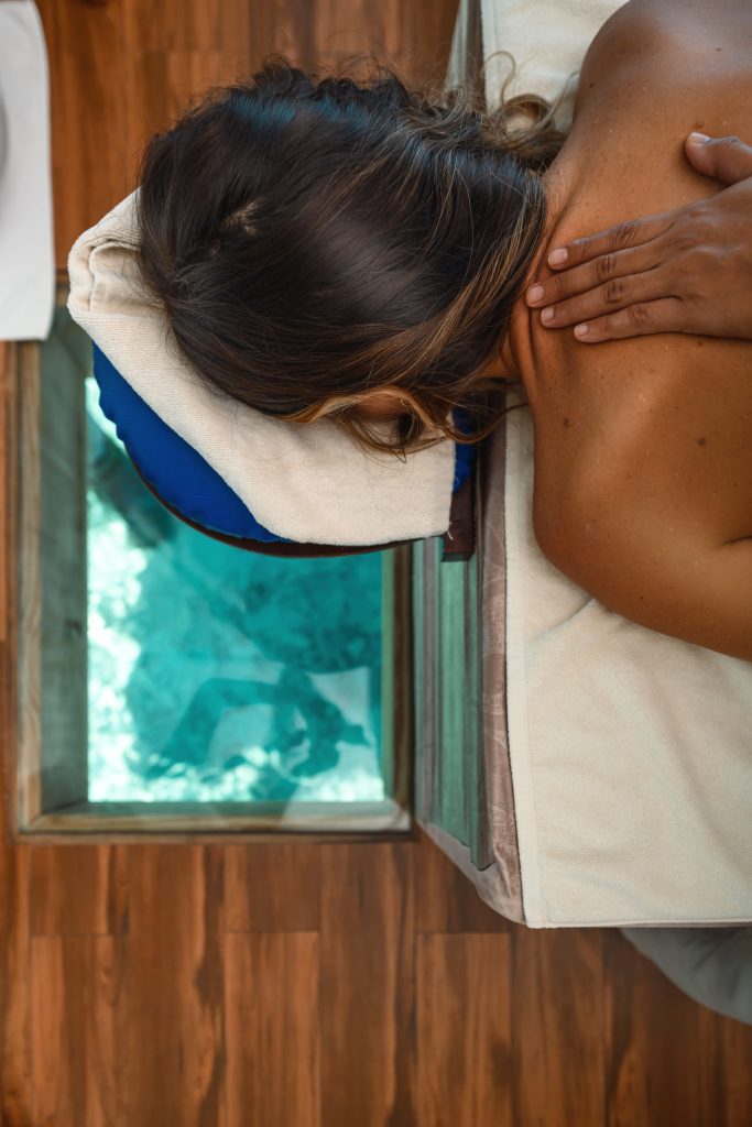 Frau liegt mit dem Gesicht nach unten auf einer Massageliege über einem Glasboden mit Blick auf Wasser