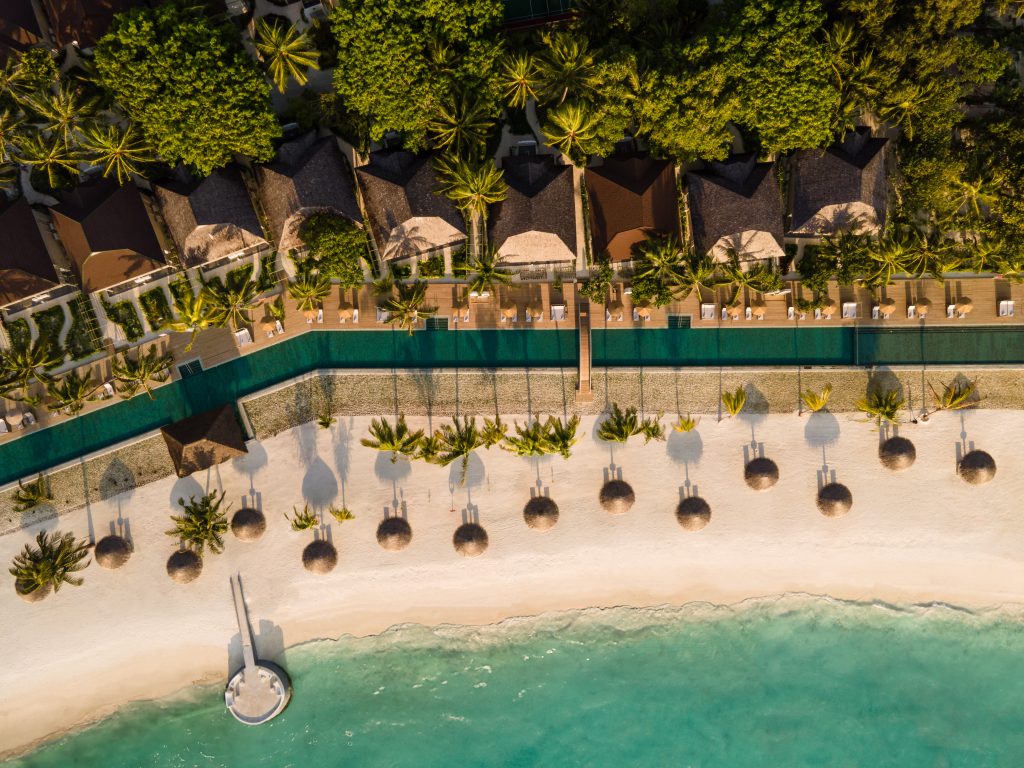 Luftaufnahme eines Strandes mit Palmen, Sonnenschirmen aus Stroh, einem Steg und Bungalows im Hintergrund