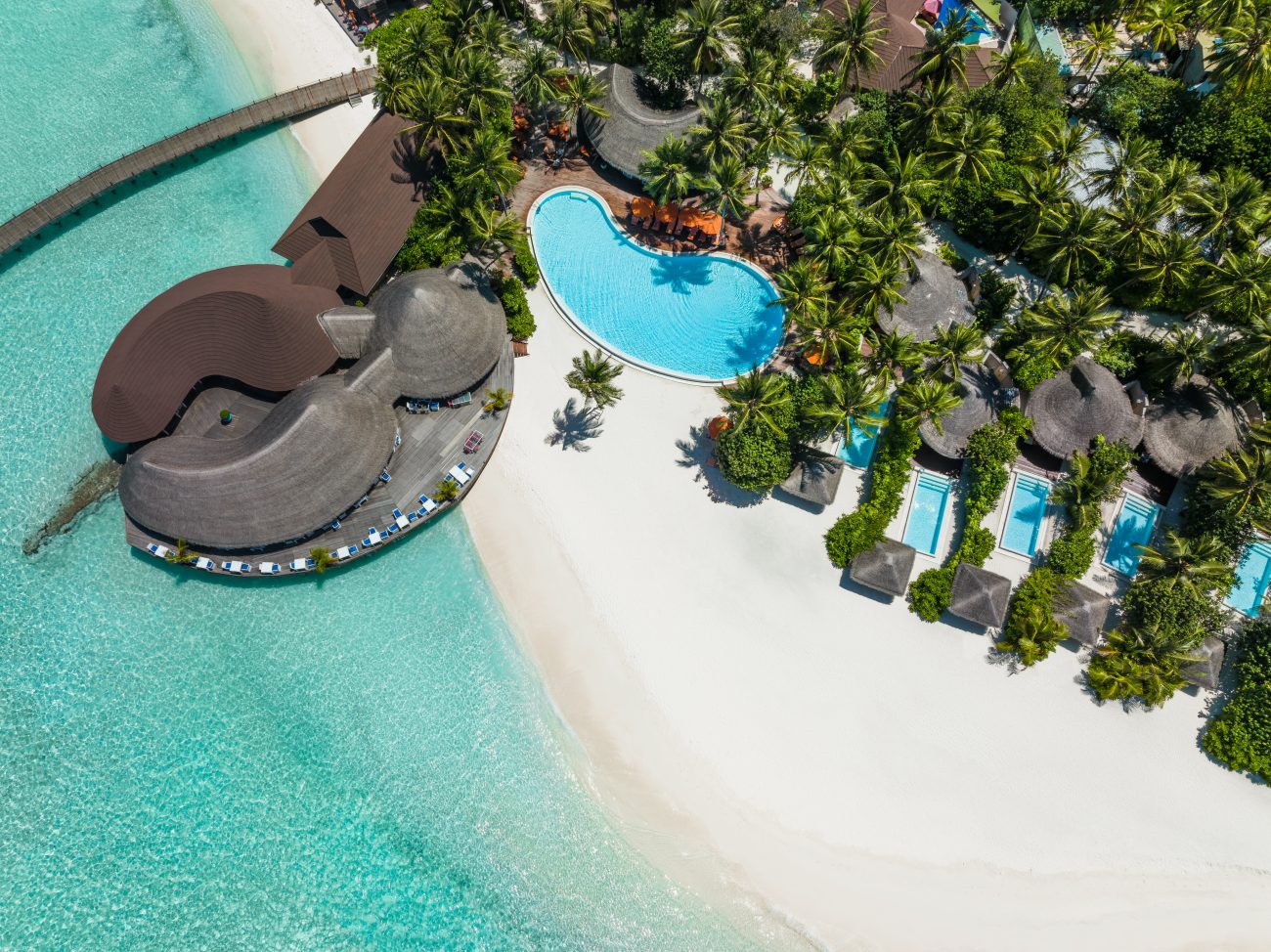 Luftaufnahme einer tropischen Strandanlage mit Pool, Palmen, Sandstrand und über Wasser gebauten Hütten