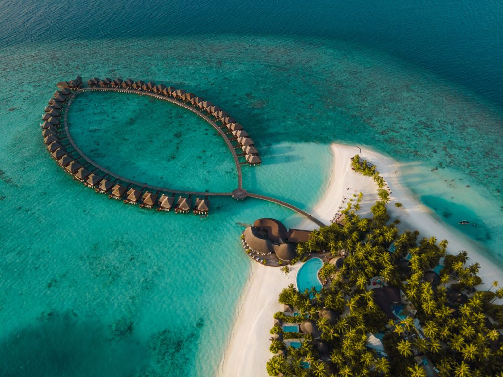 Luftaufnahme einer tropischen Insel mit Palmen, einem Pool und über dem Wasser gebauten Bungalows in einer halbkreisförmigen Anordnung