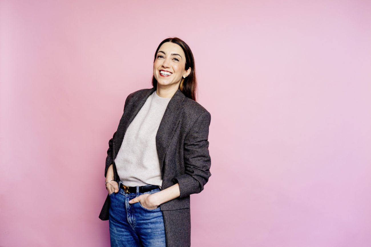 Viyan Sido mit langen dunklen Haaren lächelnd vor rosa Hintergrund, trägt grauen Pullover, dunkelgrauen Blazer und blaue Jeans