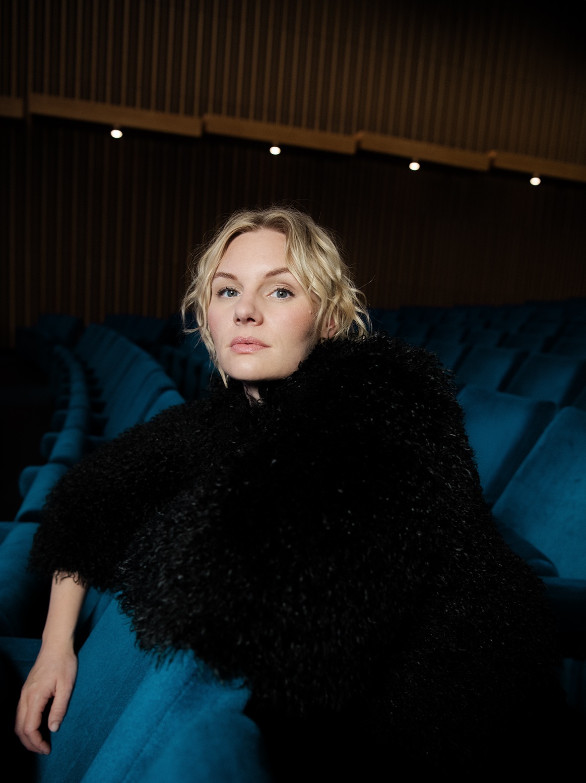 Rosalie Thomass mit blonden Haaren lehnt entspannt in einem blauen Sitz in einem dunklen Kinosaal oder Theater