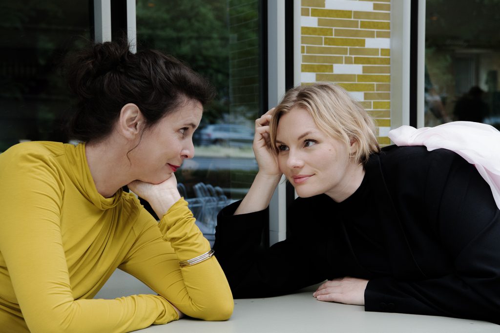 Daniela Dröscher und Rosalie Thomass sitzen sich an einem Tisch gegenüber und blicken sich an, Dröscher trägt ein gelbes Oberteil, Thomass ein schwarzes