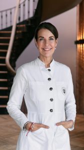 Dr. Petra Algenstaedt in weißem Arztkittel mit Emblem auf der Brusttasche steht vor einer Treppe und lächelt