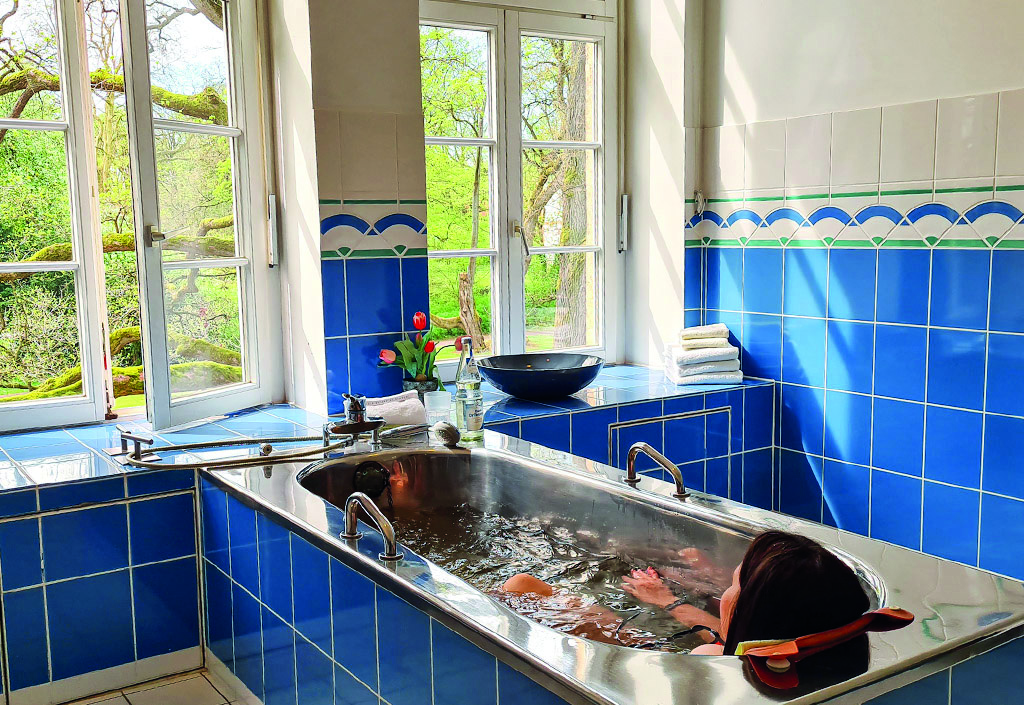 Frau liegt in einem mit blauen Fliesen verkleideten Edelstahlbad mit offenem Fenster und Blick auf grüne Bäume