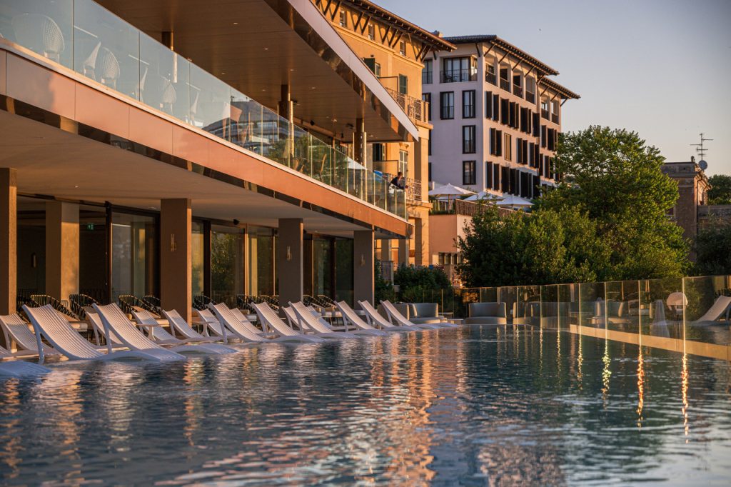 Außenpool mit Liegestühlen vor einem mehrstöckigen Hotelgebäude bei Sonnenuntergang